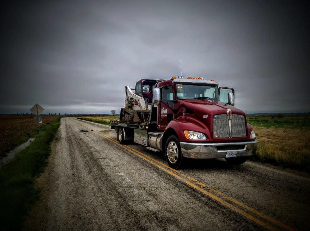 Parsons flatbed hauling a skid steer on a rural road under a storm sky